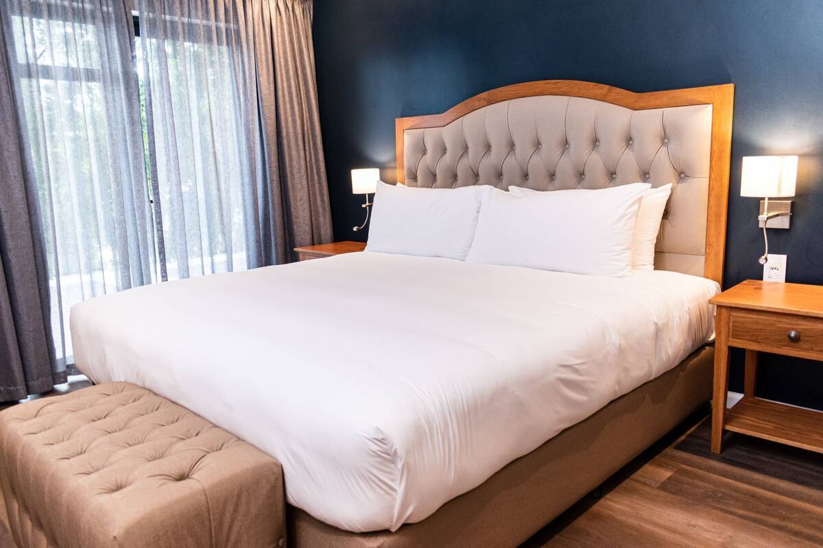 A neatly made bed with a wooden headboard, a bedside table with a lamp, and a window with curtains.