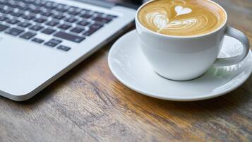 Uma chávena de café branco com arte de latte num pires ao lado de um computador portátil sobre uma mesa de madeira.
