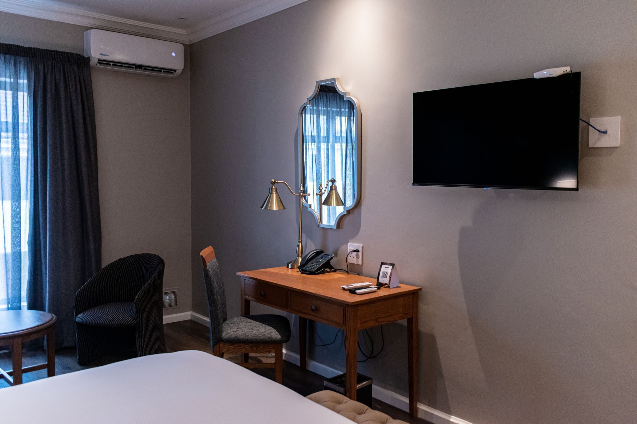 A hotel room with a bed, a desk with a telephone and a lamp, a chair, a TV mounted on the wall, and an air conditioner.