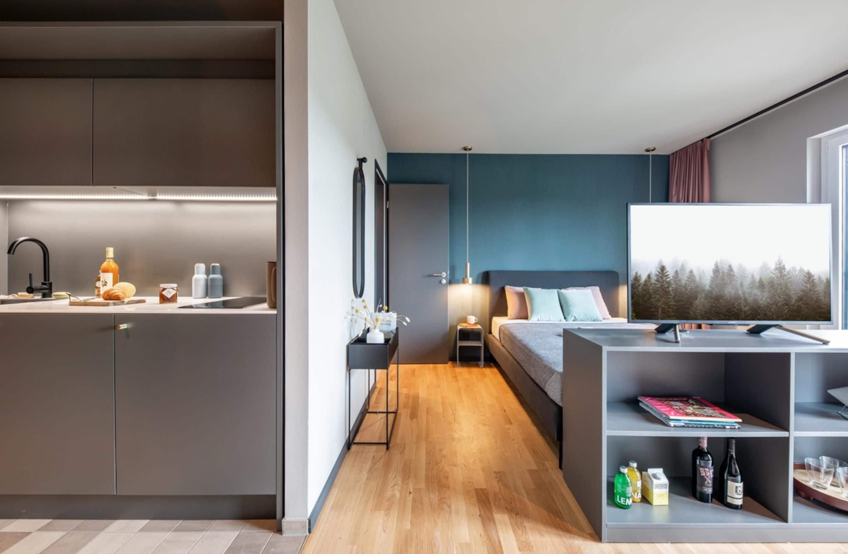 A modern hotel room with a kitchenette, a bed with a TV, and a view of trees.