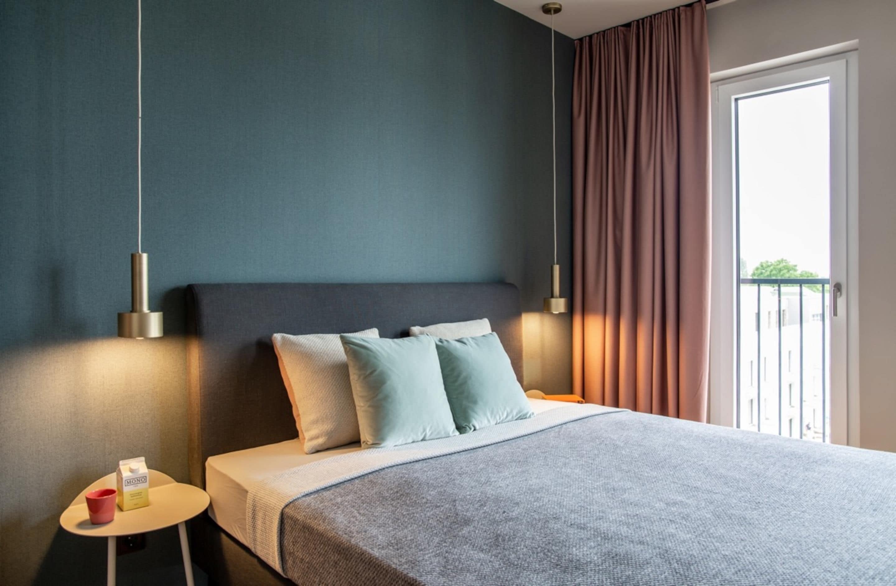 A modern hotel room with a large bed, a bedside table with a cup and a box, and a view of the outside balcony.