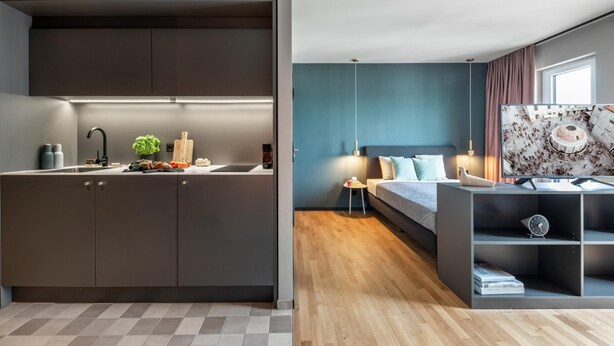 A modern kitchen with dark cabinets and a wooden countertop, a bedroom with a bed, bedside table, and a large window. - ipartment Braunschweig @HOME by Best Western (Braunschweig)