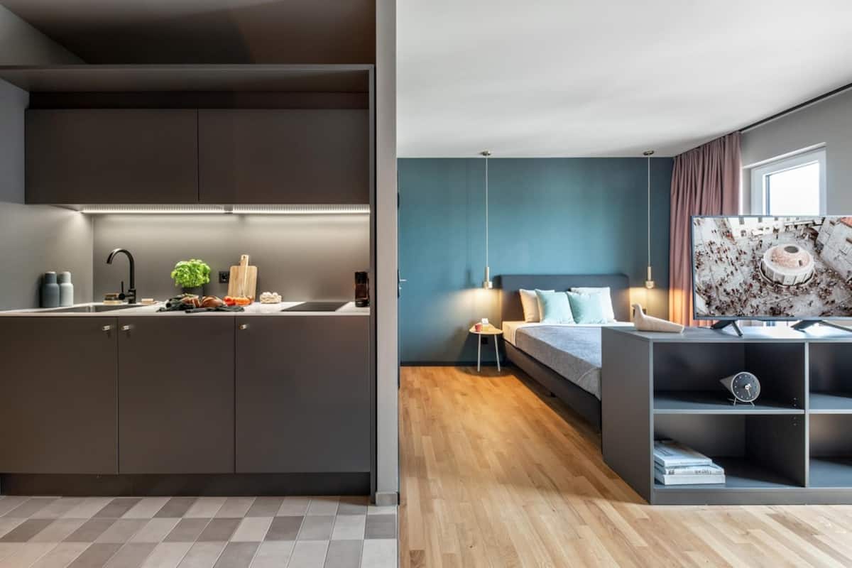 A modern kitchen with dark cabinets and a wooden countertop, a bedroom with a bed, bedside table, and a large window.