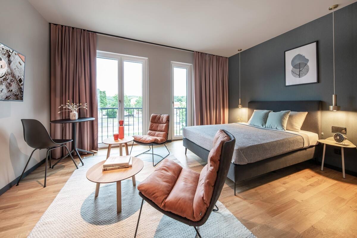 A modern bedroom with a bed, two chairs, a small table, and a balcony view.