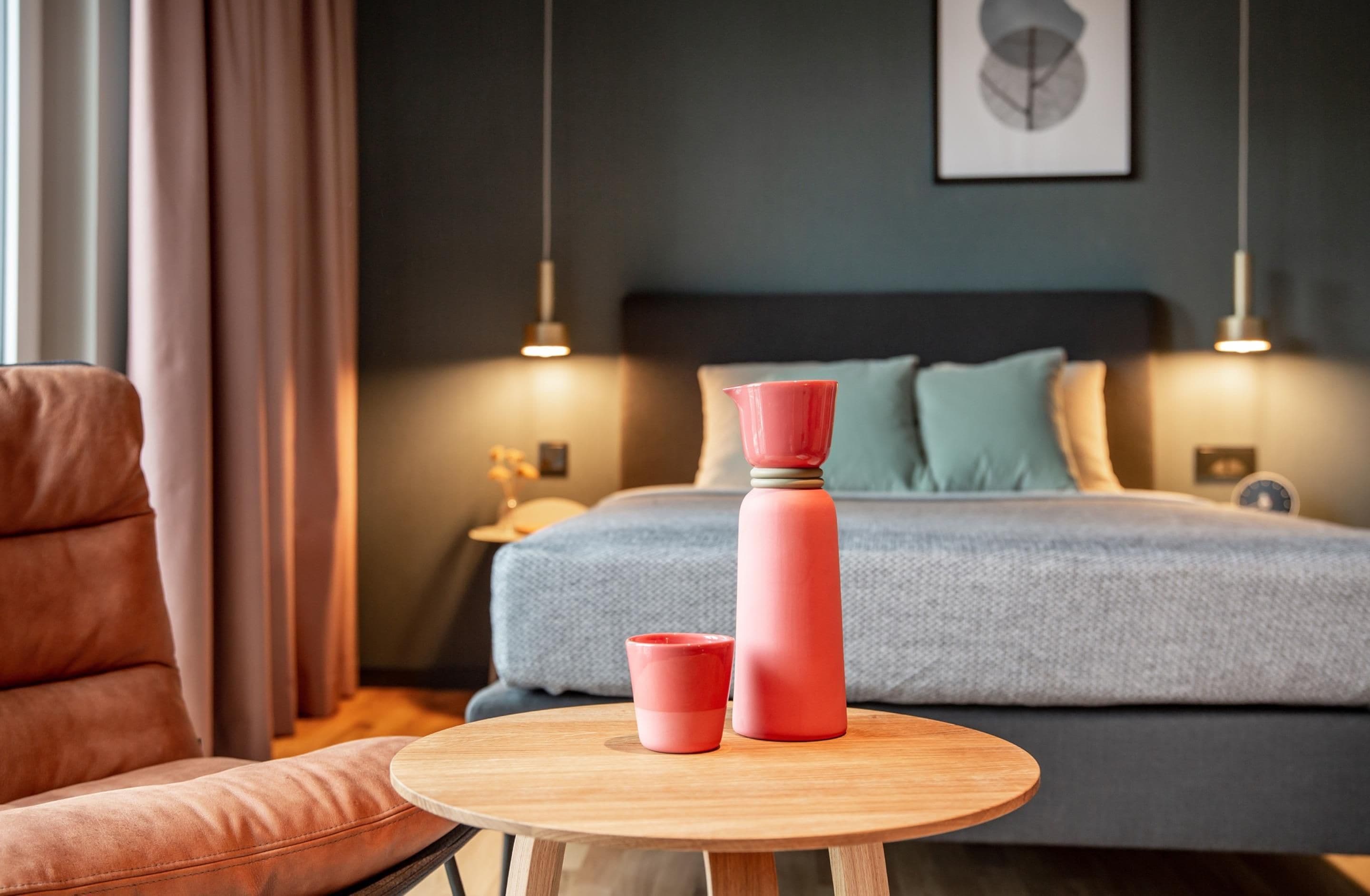 Ein Schlafzimmer mit einem Bett, Nachttischlampen, einem Nachttisch, einem Holztisch mit zwei pinkfarbenen Tassen und einem Stuhl.