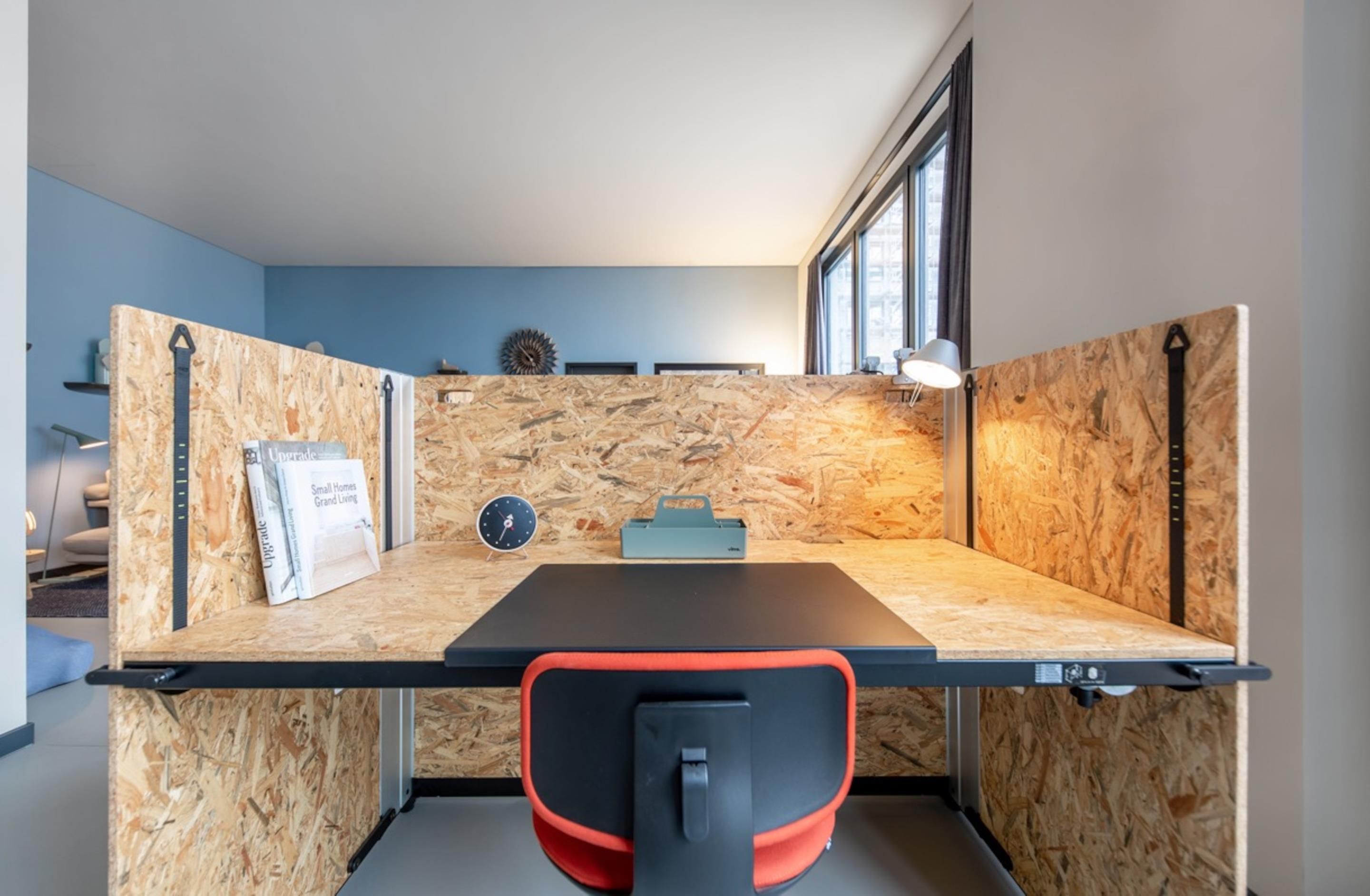 Un espacio de oficina pequeño y moderno con un escritorio de madera, un reloj, una planta pequeña y un ventanal con vistas.