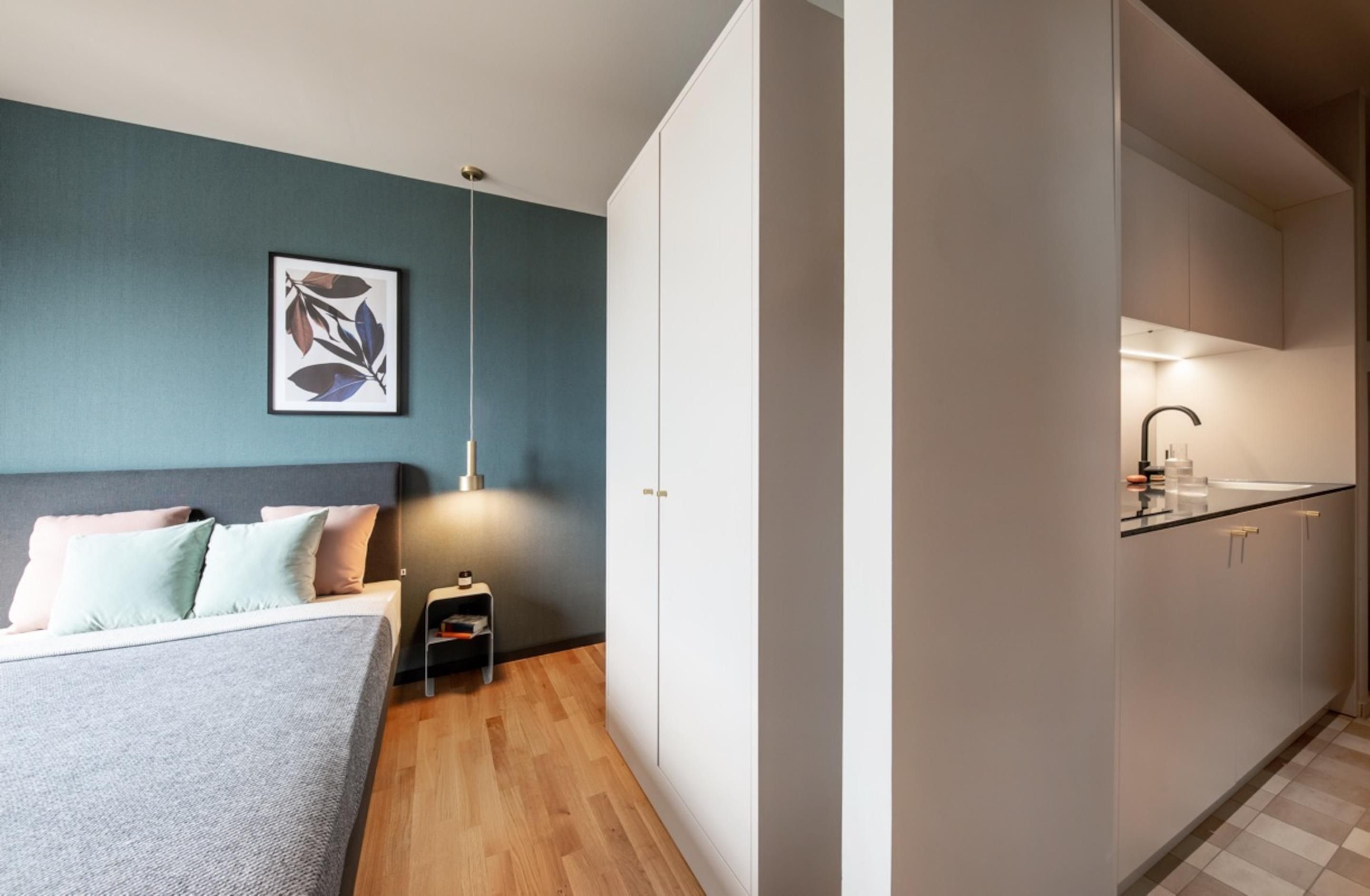Quarto compacto com cama, mesa de cabeceira e pequena mesinha de apoio. Um kitchenette adjacente inclui pia e armários.
