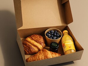 An open cardboard box containing croissants, a bowl of blueberries, a jar of jam, and a bottle of orange juice. - ipartment Braunschweig @HOME by Best Western (Braunschweig)