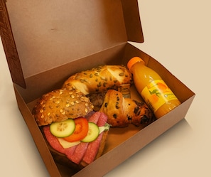 An open box containing a sandwich, a croissant, sliced vegetables, and a bottle of juice. - ipartment Braunschweig @HOME by Best Western (Braunschweig)