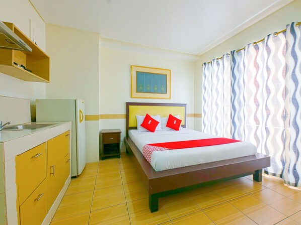 A hotel room with a bed, a small bedside table, a nightstand, a refrigerator, a sink, and a window with curtains.