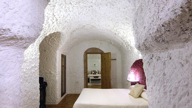 Un dormitorio acogedor de arcos, con suelo de madera, una cama con colcha blanca y una mesita de noche.