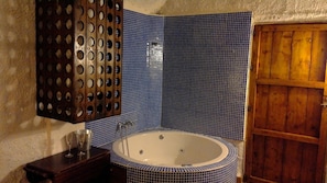 A bathroom with a large bathtub, a wooden door, and a wall-mounted sink with a metal basin. - Cuevas La Tala (Guadix)