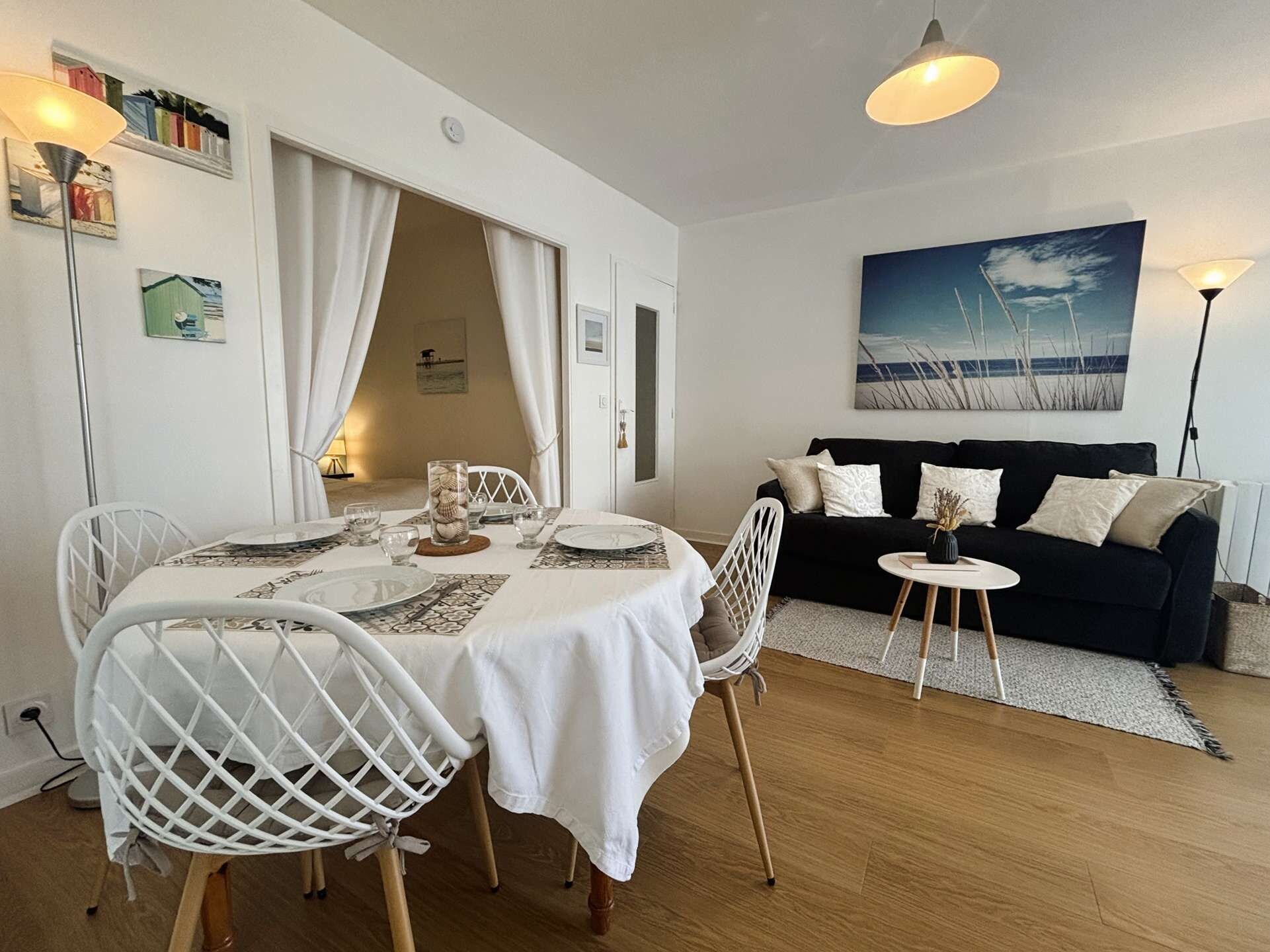 Et spiseområde med et rundt bord dækket til fire personer, hvide stole og en sort sofa i baggrunden.