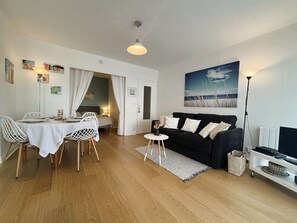 A modern living room with a dining area, a black sofa, a painting of a beach, and a TV stand.