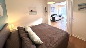 Un dormitorio con una cama grande, una mesita de noche, una lámpara y vistas a una sala de estar a través de una puerta abierta.