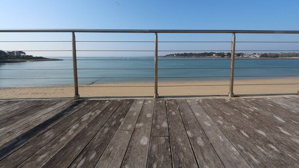 Una terraza de madera con barandilla metálica que da a una playa y un mar tranquilo.