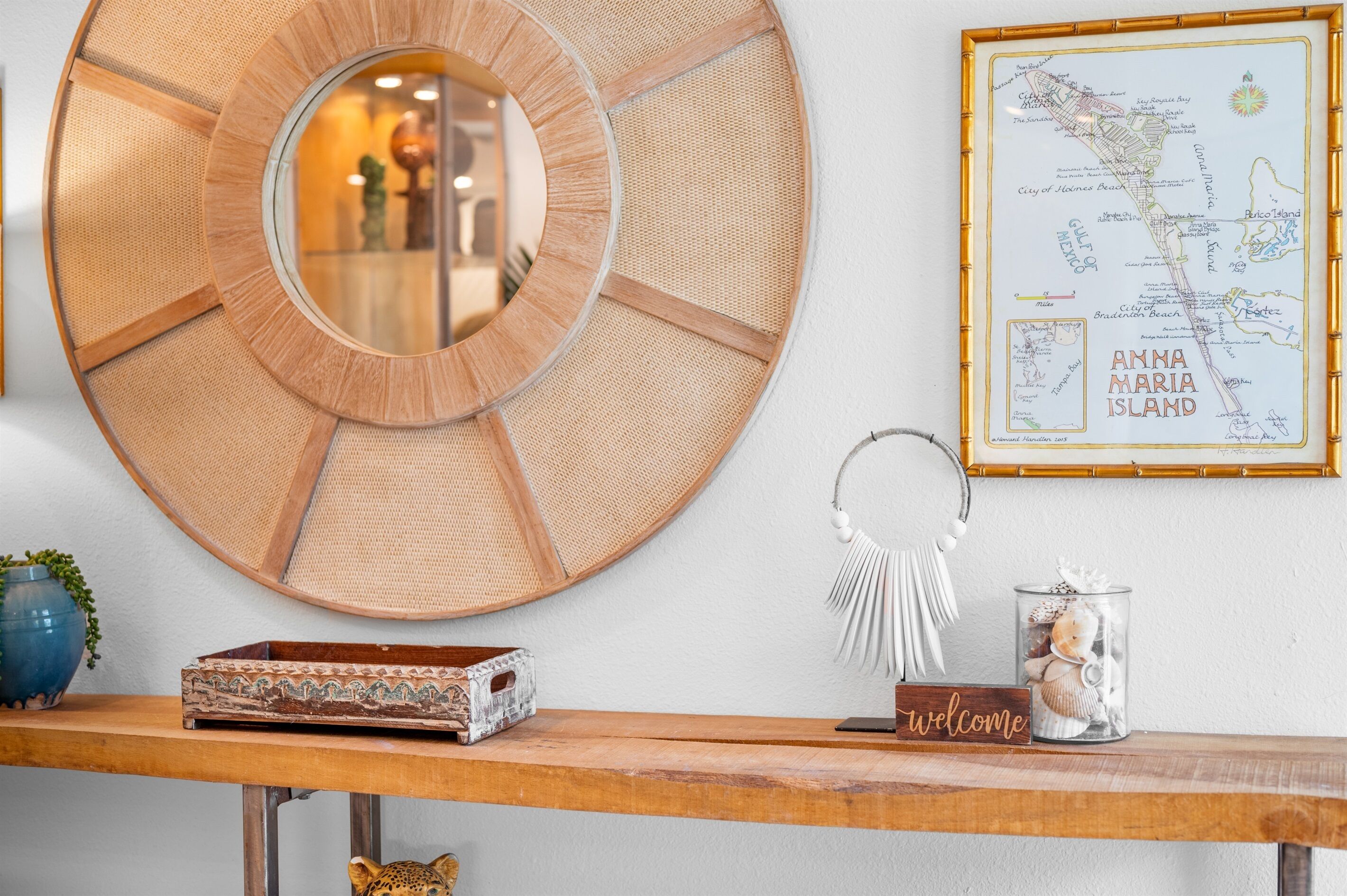 A wall with a large circular mirror, a framed map of Anna Maria Island, a welcome sign, and a decorative item with seashells.