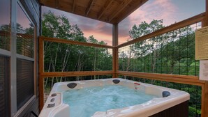 Een hottub op een houten terras met uitzicht op bomen en een zonsondergang.