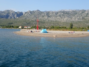 Una playa con tiendas de campaña y gente tomando el sol, una torre roja y montañas al fondo.