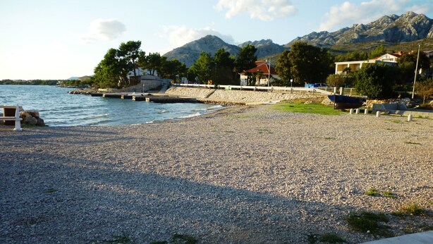 Una playa de guijarros con un muro de piedra, un muelle y edificios al fondo.