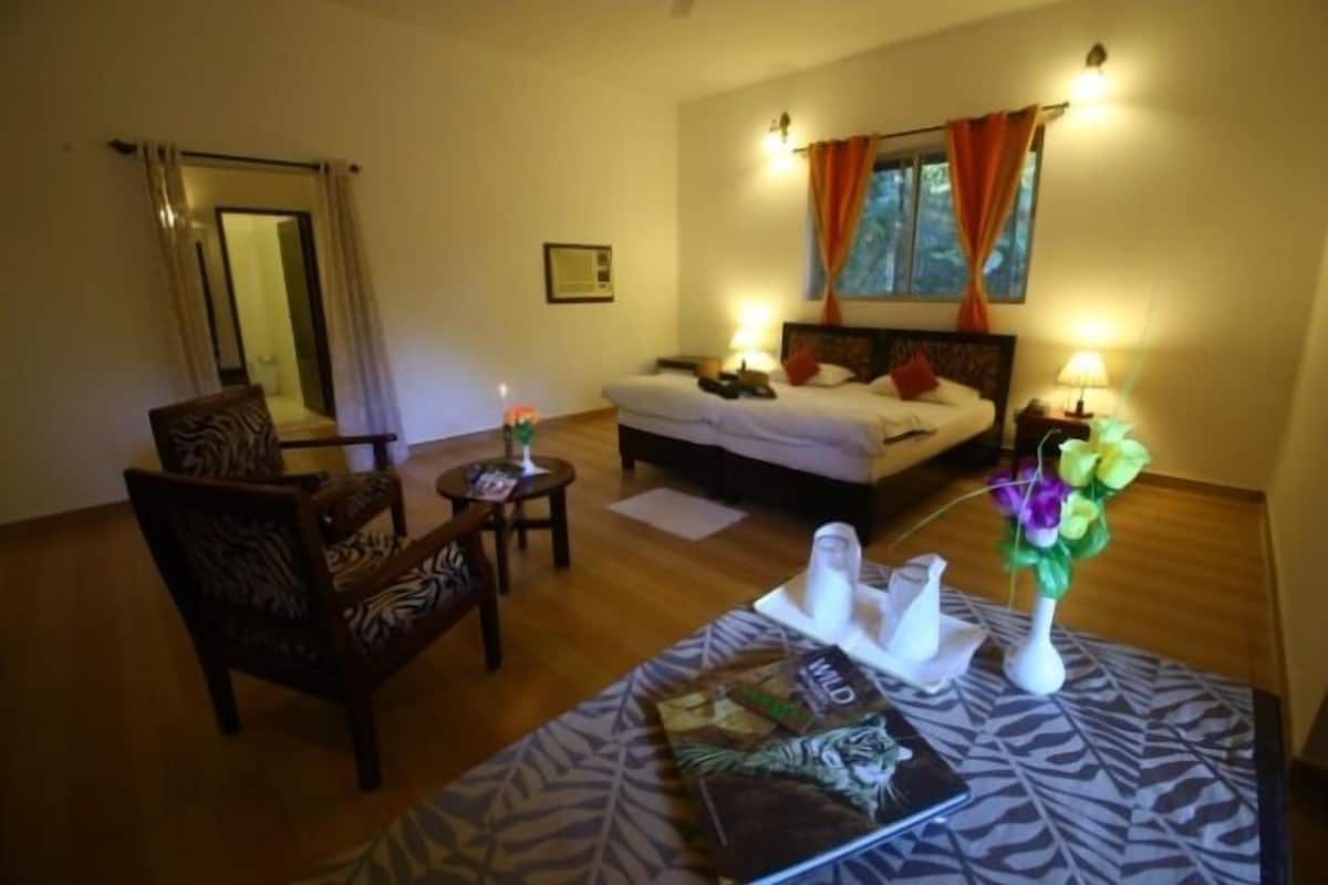 Una habitación de hotel con una cama, una silla, una mesita con una revista y un florero con flores.
