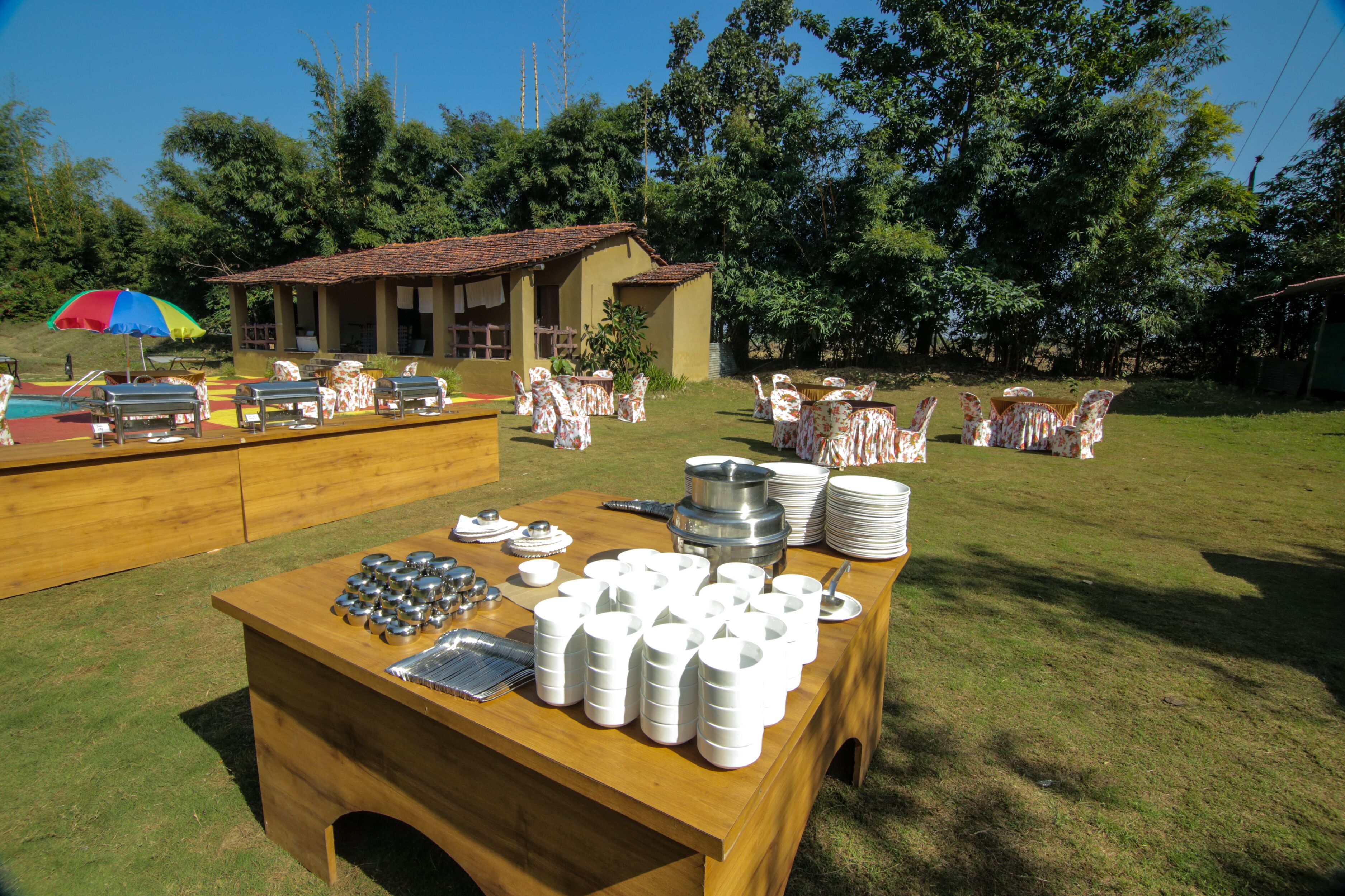 A buffet setup with a wooden table, white plates, and silverware, surrounded by chairs and a small building in a grassy area.