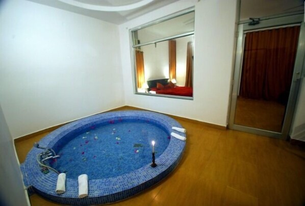 Habitación con un pequeño jacuzzi de azulejos azules, suelo de madera, un espejo grande que refleja una cama con ropa de cama roja y una puerta corredera con cortina.