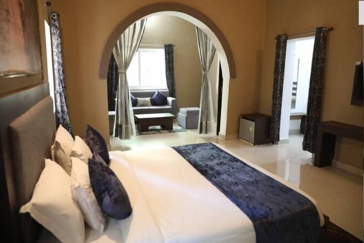 Un dormitorio con una cama grande, un arco que da a una sala de estar con un sofá y una mesa, y un televisor en la pared.