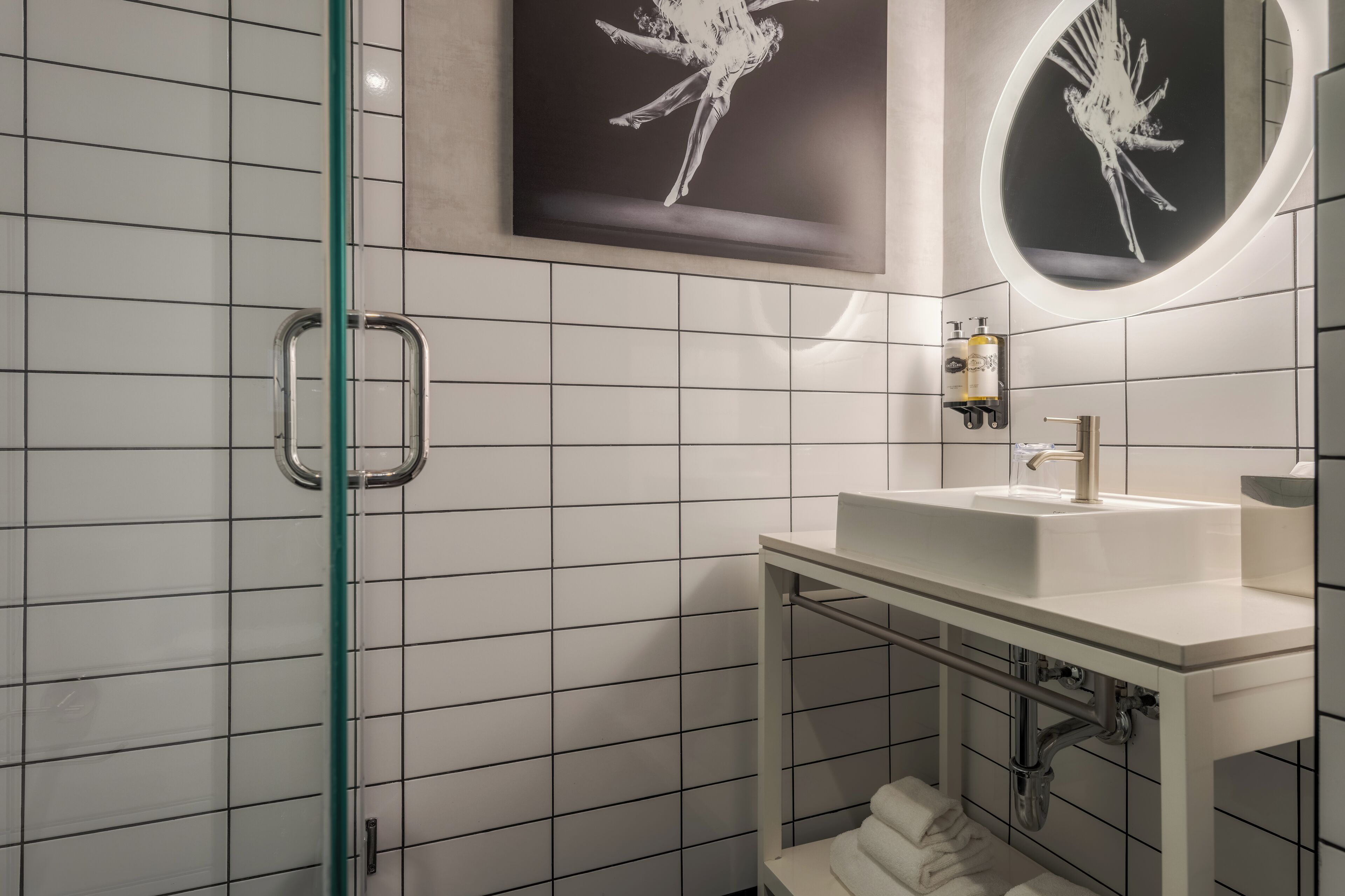 A modern bathroom with white tiled walls, a glass shower enclosure, a white sink with a mirror, and two black-and-white artwork pieces.