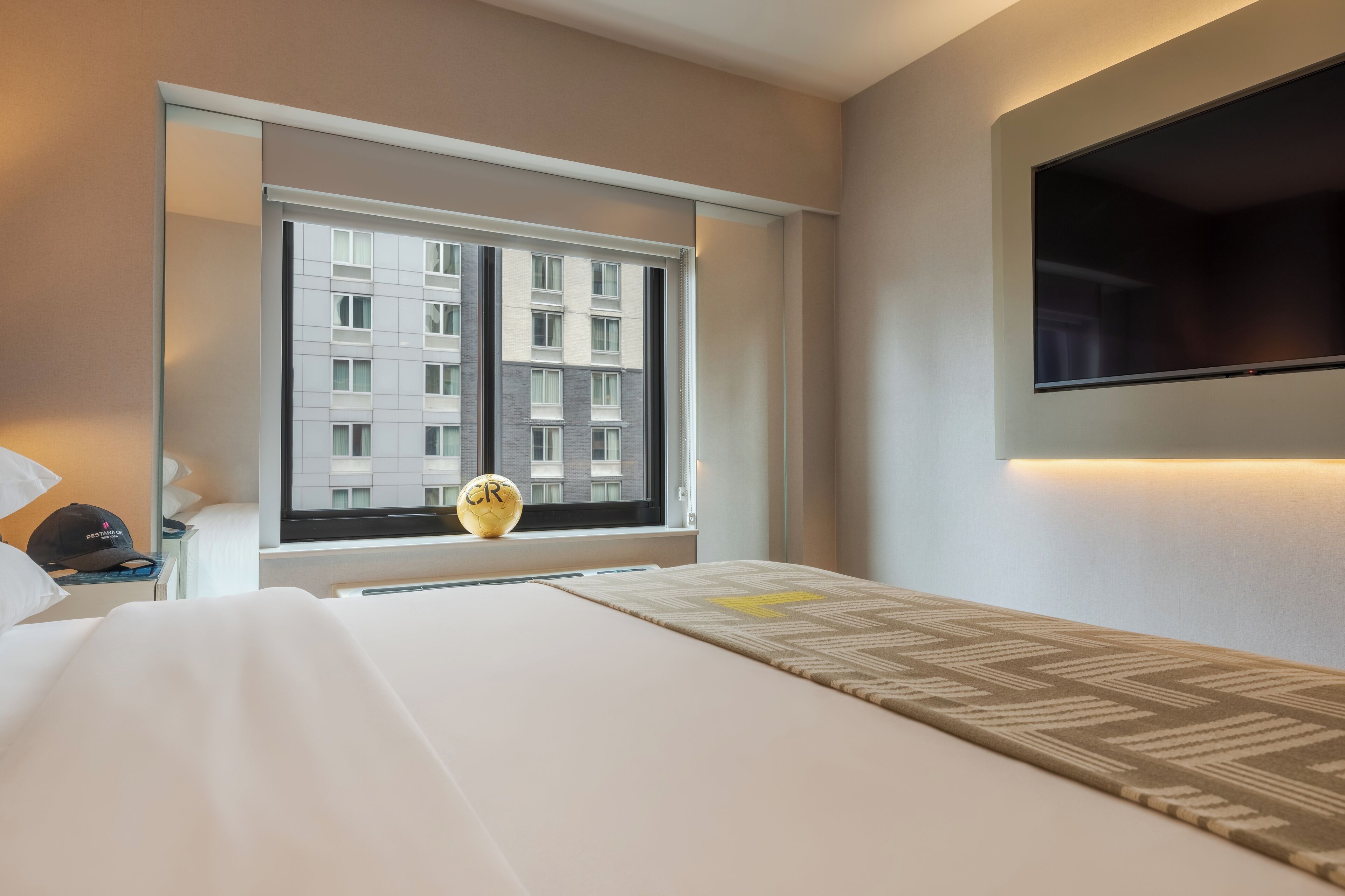 Quarto de hotel com uma cama, televisão na parede, janela com vista para um edifício e uma pequena mesa com um abajur e um copo.