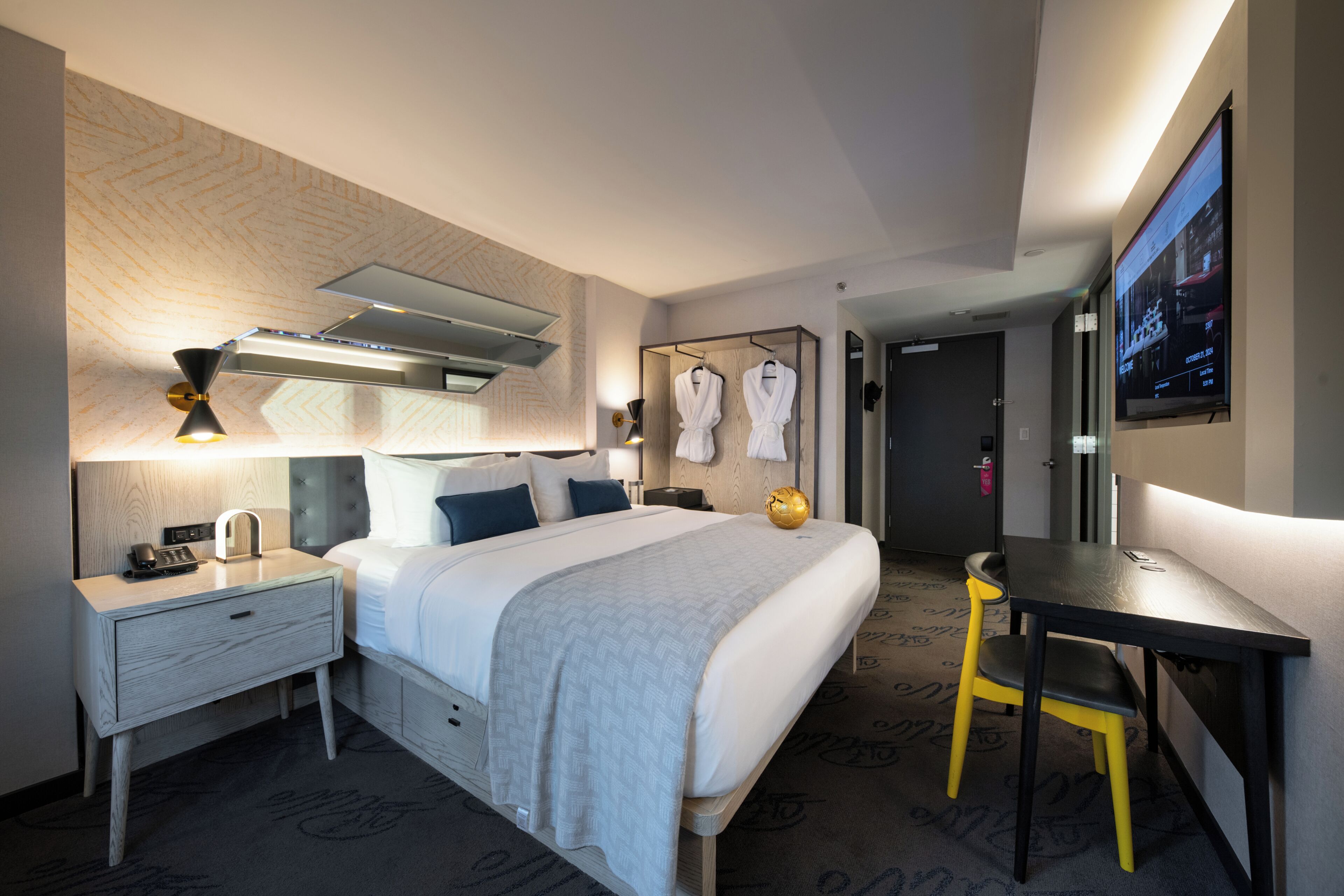 A hotel room with a bed, bedside table, lamp, TV, and a desk with a chair.