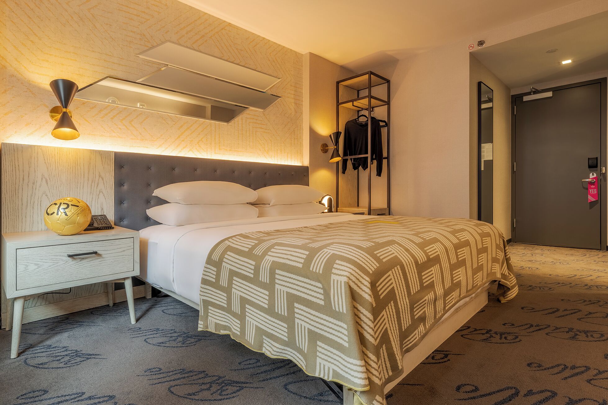Een hotelkamer met een bed, nachtkastje, lamp en een wandplank met kleding.