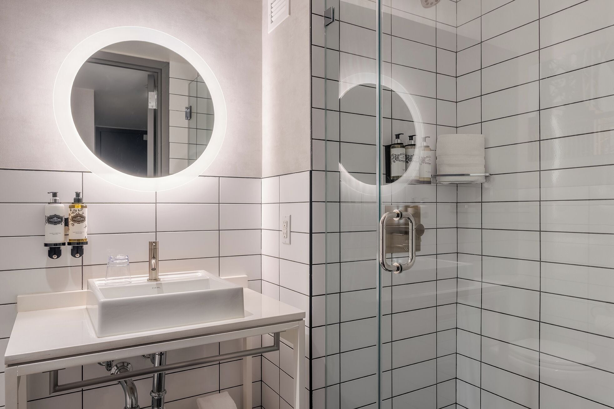 A modern bathroom with a white sink, a mirror with a circular light, and a glass shower enclosure.