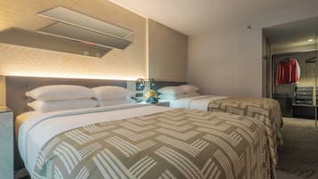 A hotel room with two beds, a headboard with built-in lighting, a bedside table with a lamp, and a wardrobe with a red shirt hanging.