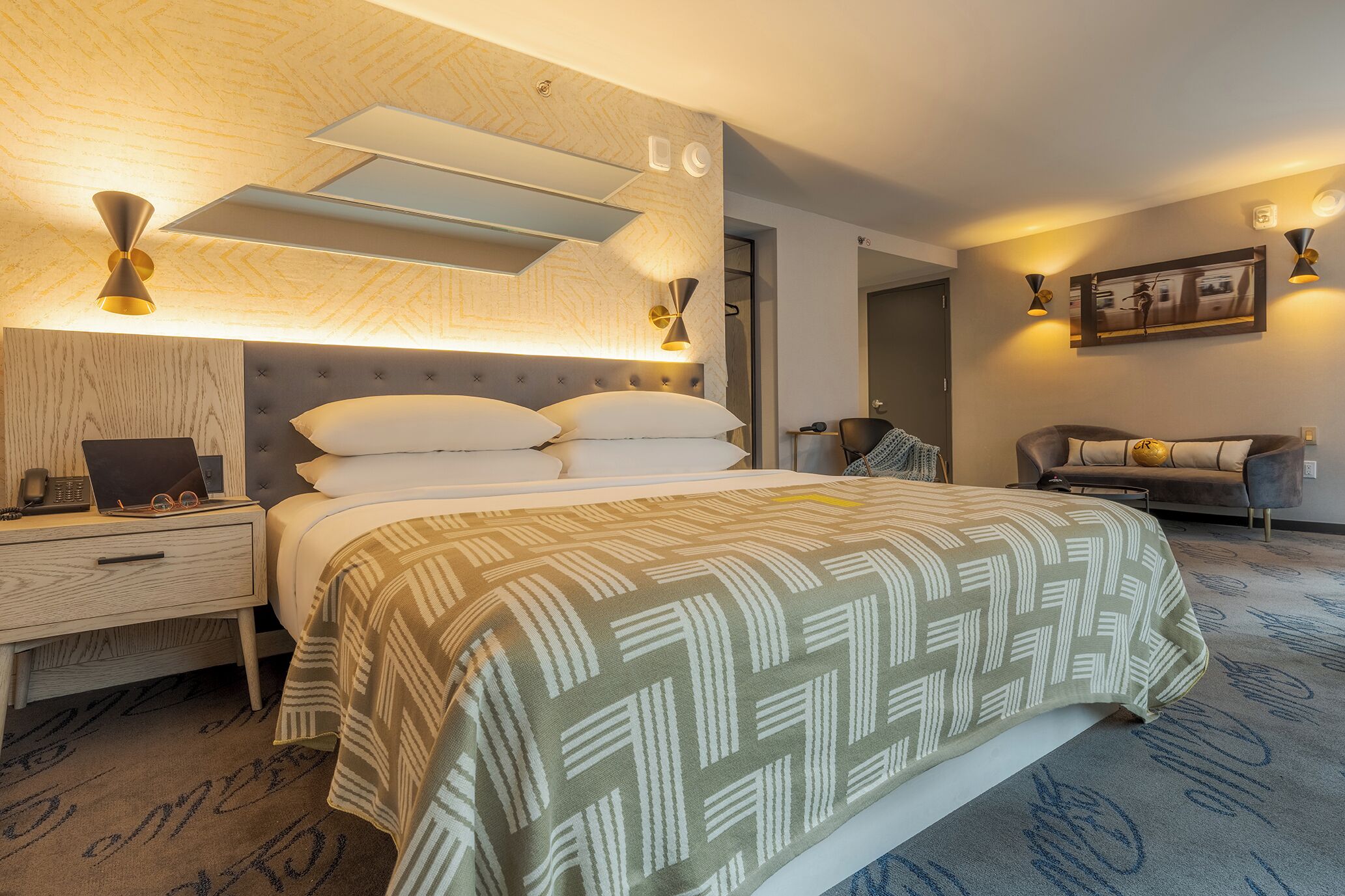 A modern hotel room with a large bed, a bedside table with a lamp, a sofa, and a wall-mounted artwork.