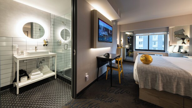 A hotel room with a bed, a desk, a chair, a TV, a bathroom with a sink and mirror, and a view of buildings outside. - Pestana CR7 Times Square (New York)