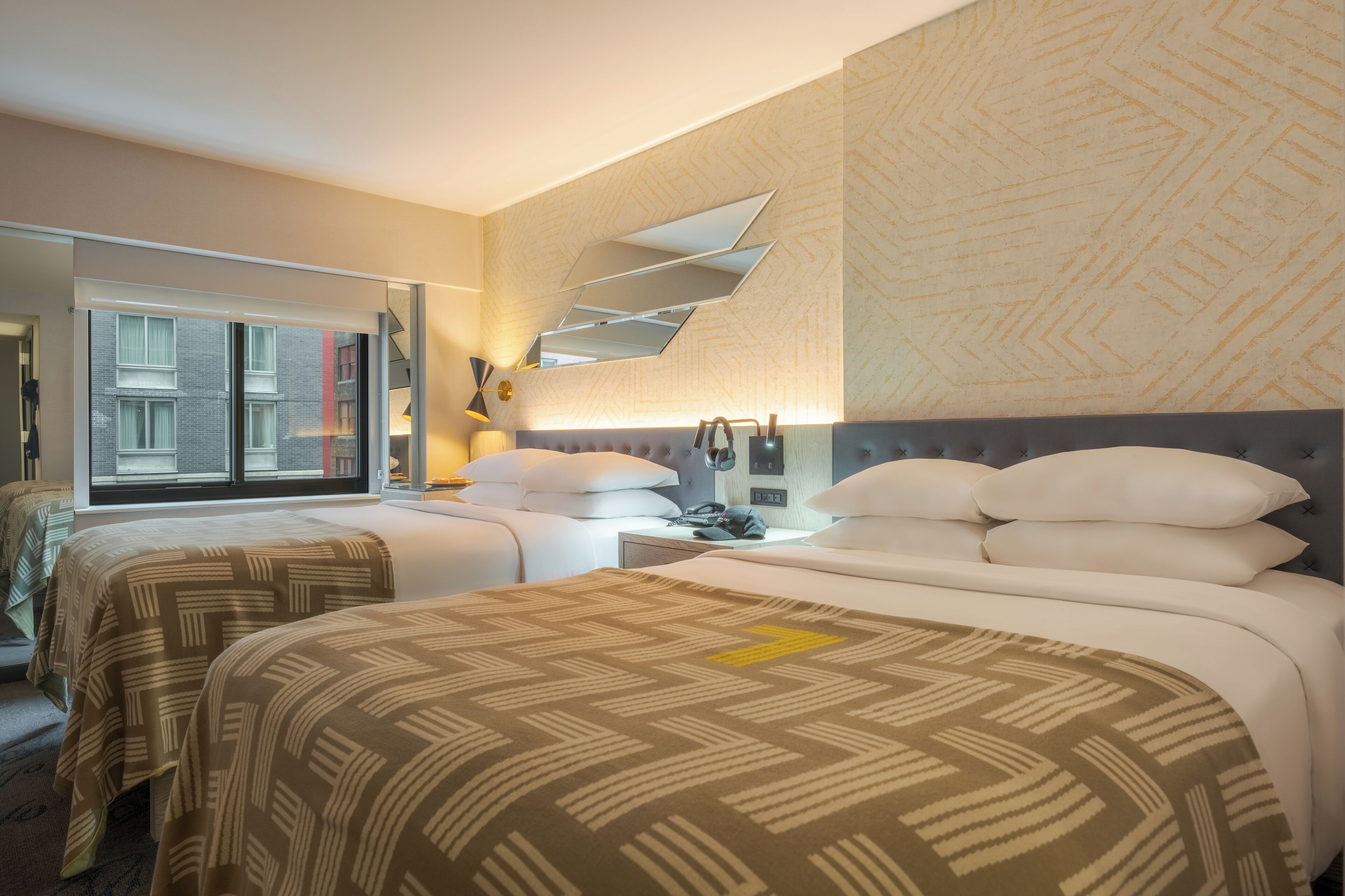 A hotel room with two beds, a large window, a mirror, and a wall with a geometric pattern.