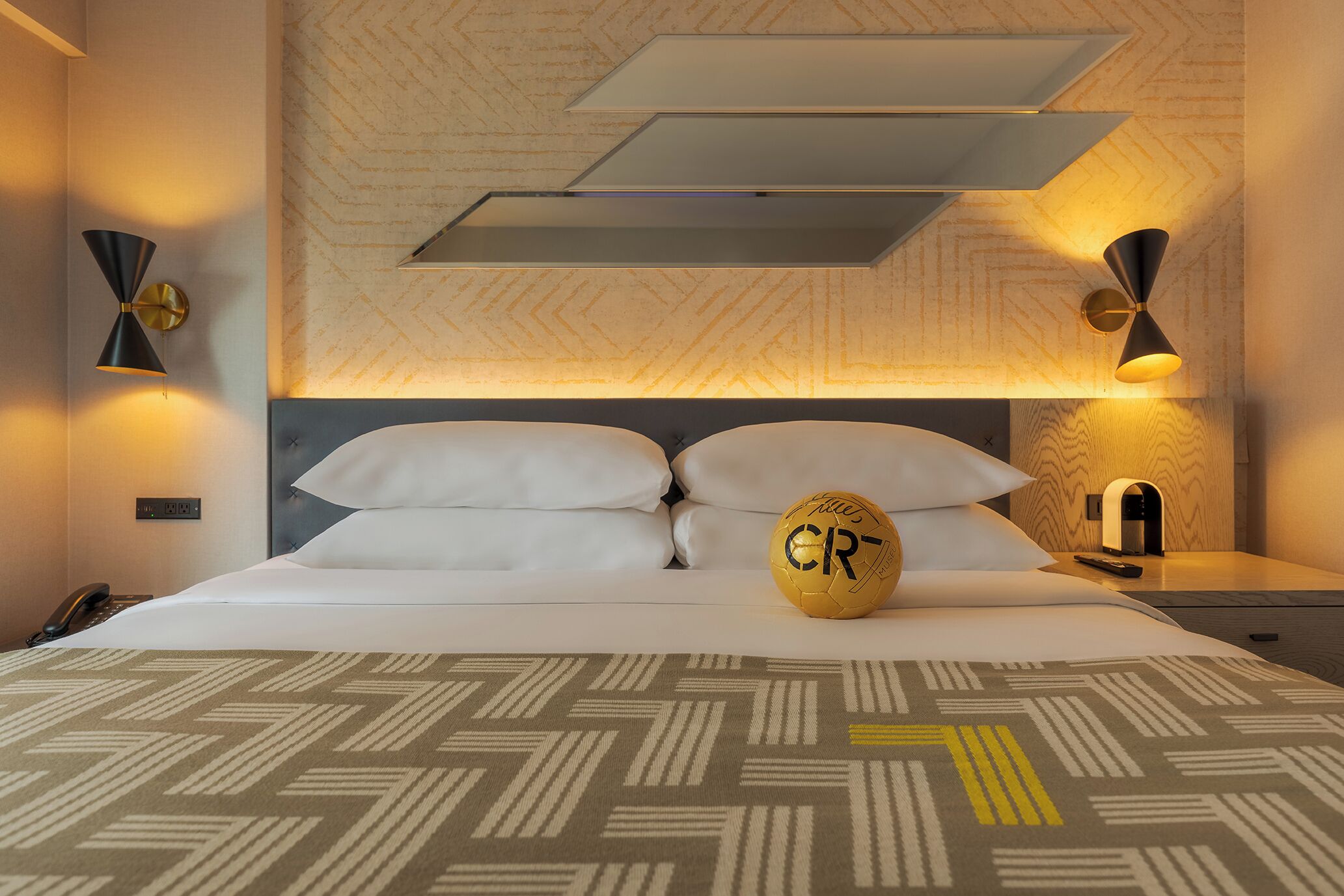 A hotel room with a bed, two bedside lamps, a bedside table, and a wall-mounted shelf.