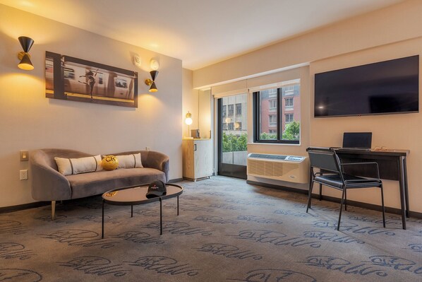 A modern hotel room with a sofa, a desk, a chair, a flat-screen TV, a painting, and a large window offering a city view.