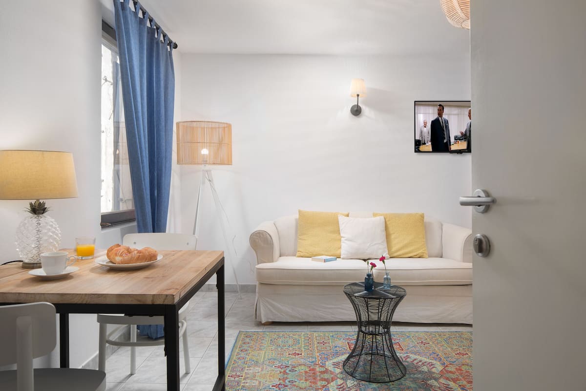 A living room with a wooden table, a white sofa, a patterned rug, and a television on the wall.