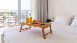 A neatly made bed with a white comforter, a wooden bedside table with breakfast items, and a view of the city through large windows. - Top Tier Apartment with Swimming Pool (Herzliya)