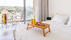 A hotel room with a bed, a wooden tray with fruits and drinks, a laptop on a nightstand, and a balcony with a table and chairs. - Top Tier Apartment with Swimming Pool (Herzliya)