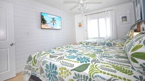 Une chambre avec des murs lambrés blancs, un ventilateur de plafond, un lit avec une literie à motifs floraux, un tableau sur le thème de la plage accroché au mur et une fenêtre avec des rideaux.