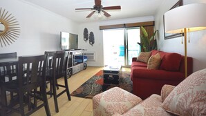 Un salon avec un canapé rouge, un ventilateur de plafond, un téléviseur et un coin repas comprenant des chaises et une table.