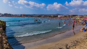 Ein Strand mit klarem, blauem Wasser, Menschen beim Schwimmen und Sonnenbaden, und Gebäuden im Hintergrund.