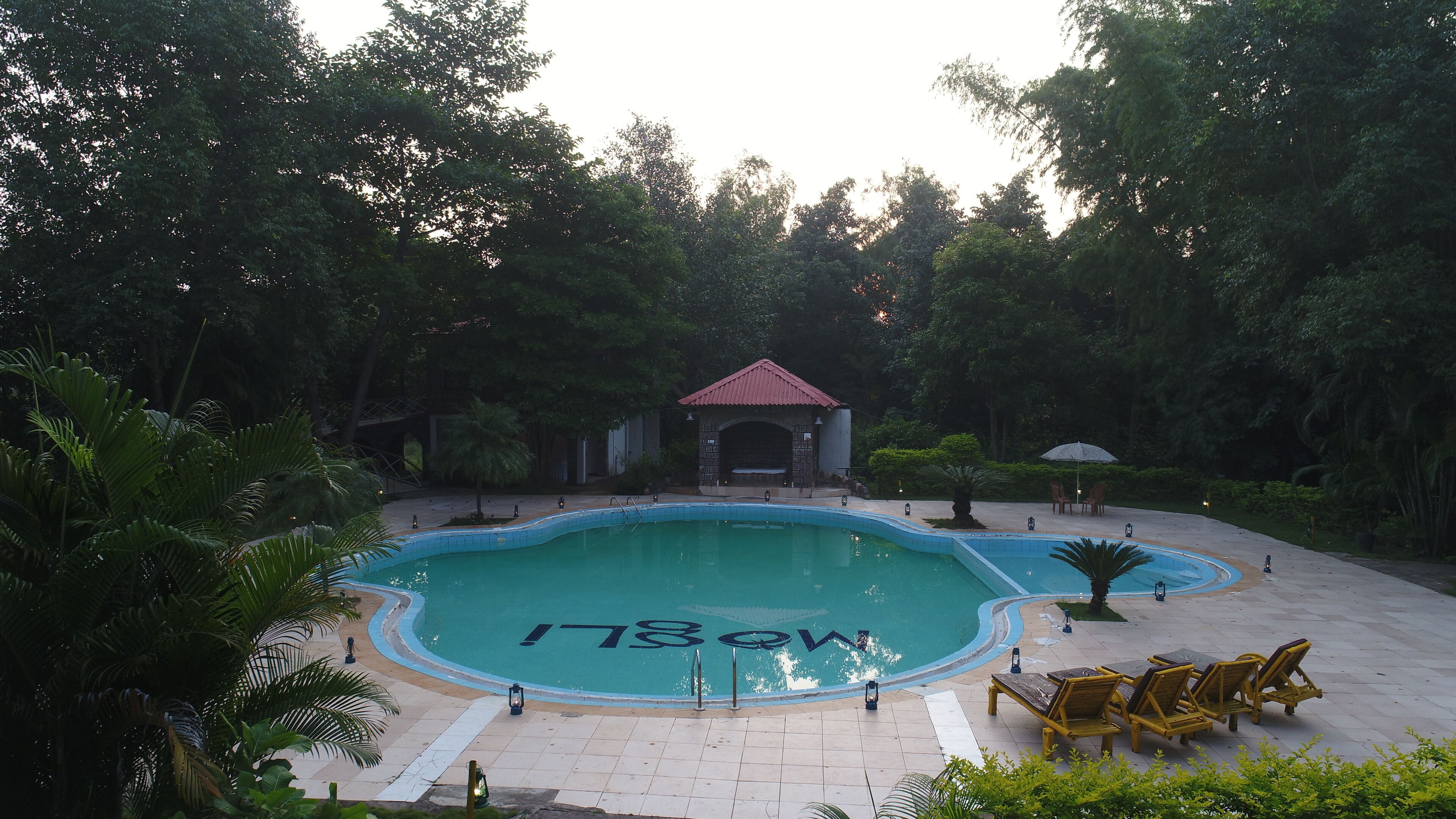 A swimming pool with a tiled deck, surrounded by lush greenery and a small pavilion.