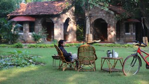 A person sitting on a chair in a garden with a bicycle and a table with a teapot and cups.