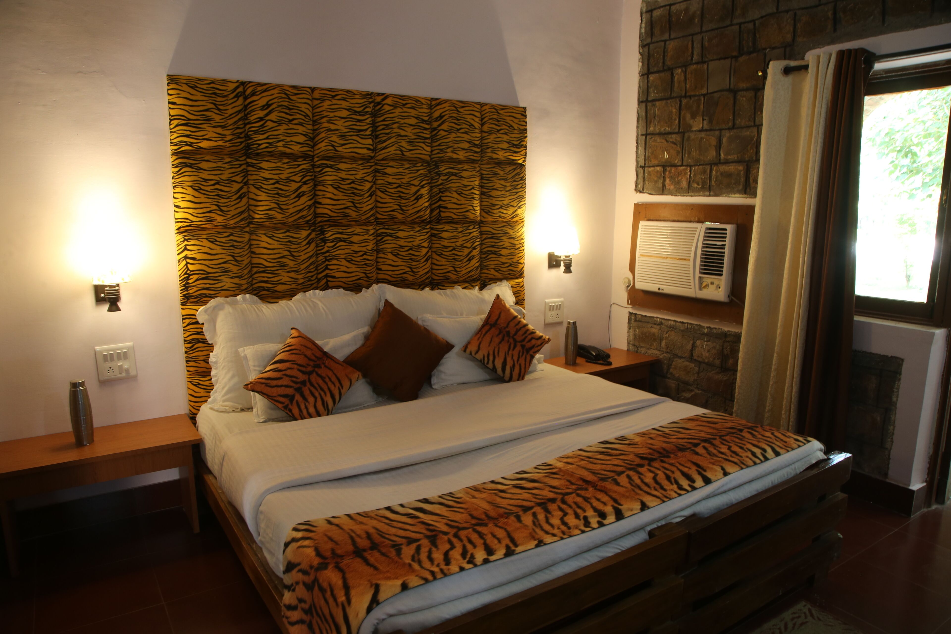 Cama com cabeceira estampada de tigre, roupa de cama branca e manta com padrão de tigre. Há duas mesas de cabeceira com luminárias e um ar condicionado fixado na parede.