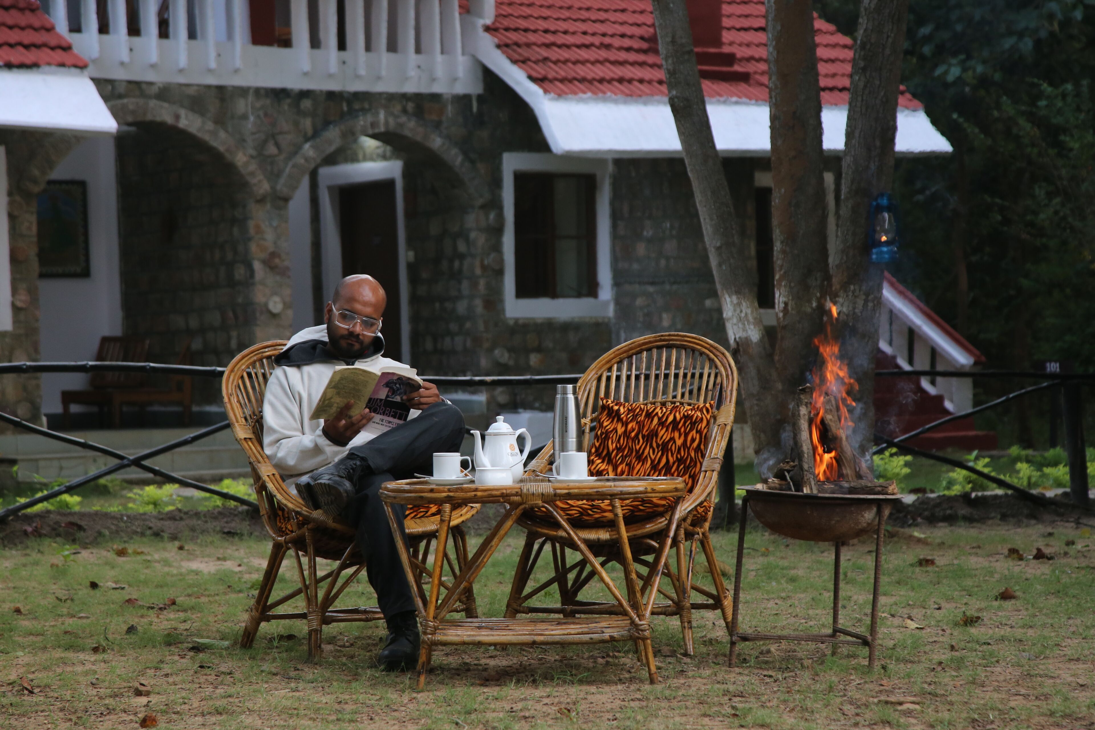 A man reading a book at a wicker table with a fire pit nearby.