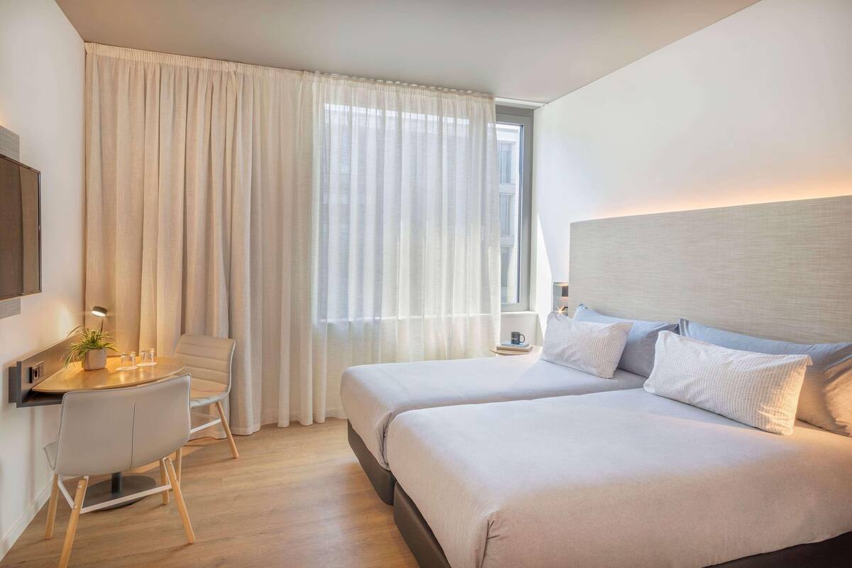 Quarto de hotel com duas camas, uma escrivaninha com cadeira, uma mesinha com uma planta e uma janela com cortinas.